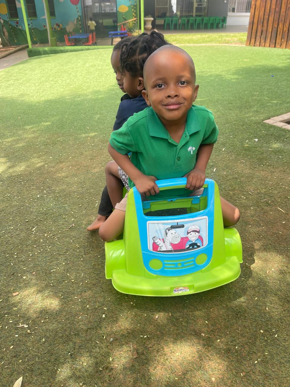 Two friends sharing a green ride-on car on the school's soft green lawn