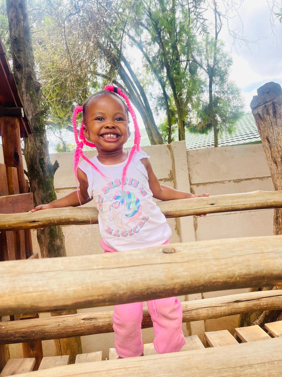 A beaming little girl with pink braids standing on the wooden jungle gym