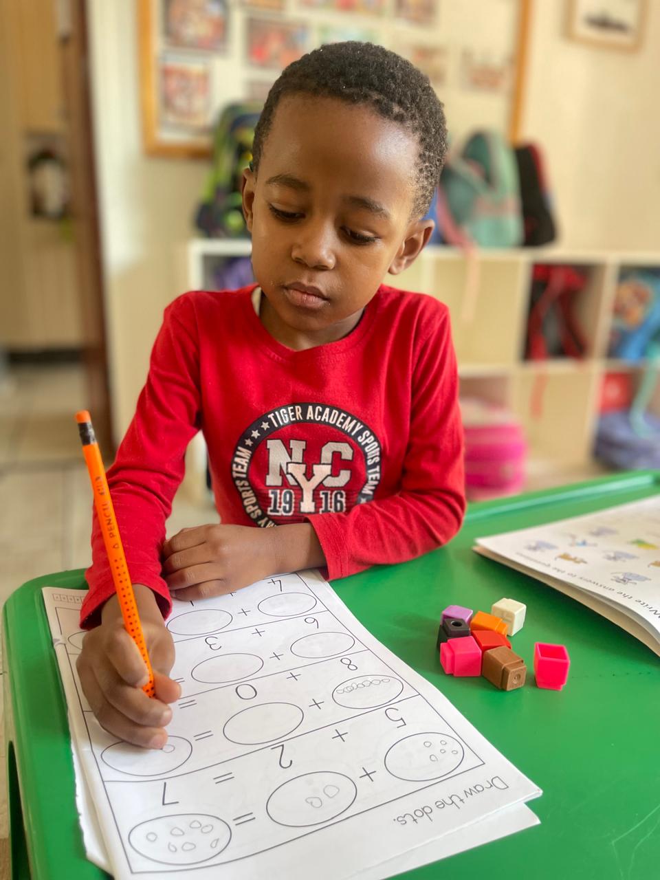 A boy in a red top concentrating on a number worksheet with counting blocks