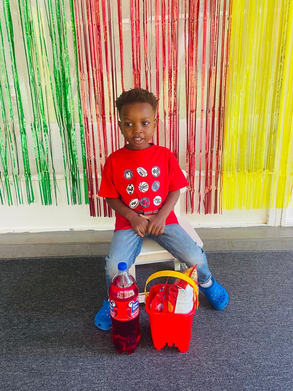 A little boy in a red sports shirt sitting in front of a bright party backdrop with his treat bucket